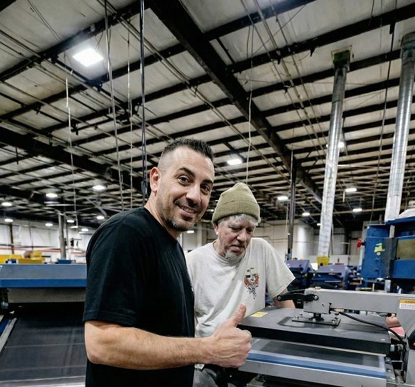 Two men at a print shop give a thumbs up beside a heat press