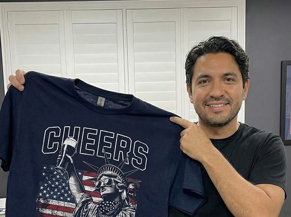 A man holds up a navy tee printed with CHEERS and the Statue of Liberty